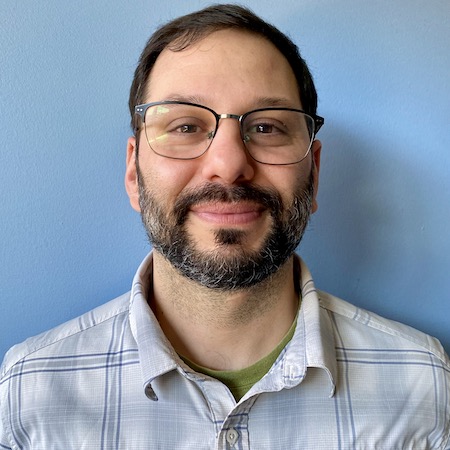 Gerasimos has short brown hair, a short brown beard with a few white patches, and light skin. He is wearing a blue and white collared shirt with a broad plaid pattern. He is smiling with his mouth closed in front of a plain sky-blue wall.