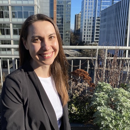 Mikala has light brown hair draped over her shoulder and light skin. She is wearing a dark gray blazer with a white shirt underneath. She smiles with her teeth showing, standing near a balcony railing and a variety of potted plants. Large city buildings cover most of the background, with a bit of blue sky in the middle.