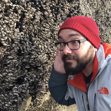 Robert has a brown beard, light skin, and his hair is covered with a bright red beanie. He wears glasses and an orange/blue/white zippered coat. He is wide-eyed and has his hand to his ear, listening to the many barnacles on the large rock right behind him.
