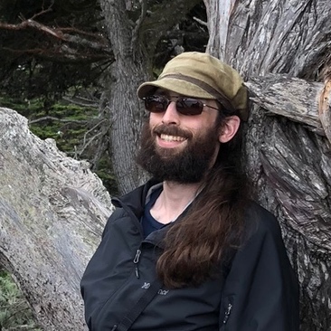Steven has long brown hair draping over his shoulder, a full brown beard, and light skin. He is wearing sunglasses, an olive green billed cap, and a black rain jacket. His teeth are showing as he smiles and leans against a tree, with a forest in the background.